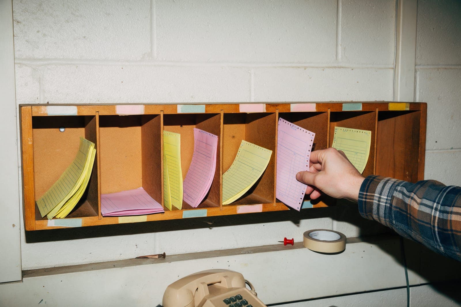 A wall of wooden cubbies holding canary and pink carbon copies, a hand pulling one pink copy out.