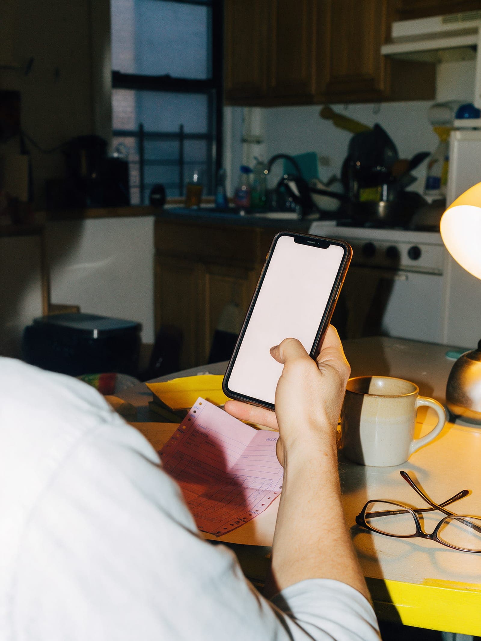 A hand holding a phone over a folded pink carbon copy on a kitchen table at night.