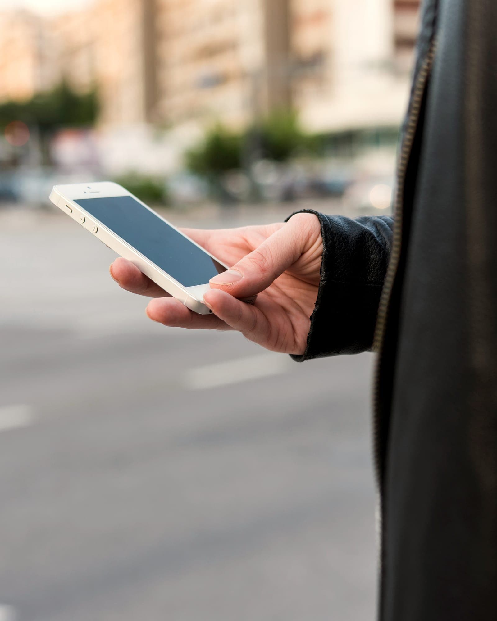 A hand holds a phone on a city street, apartment blocks blurred behind.