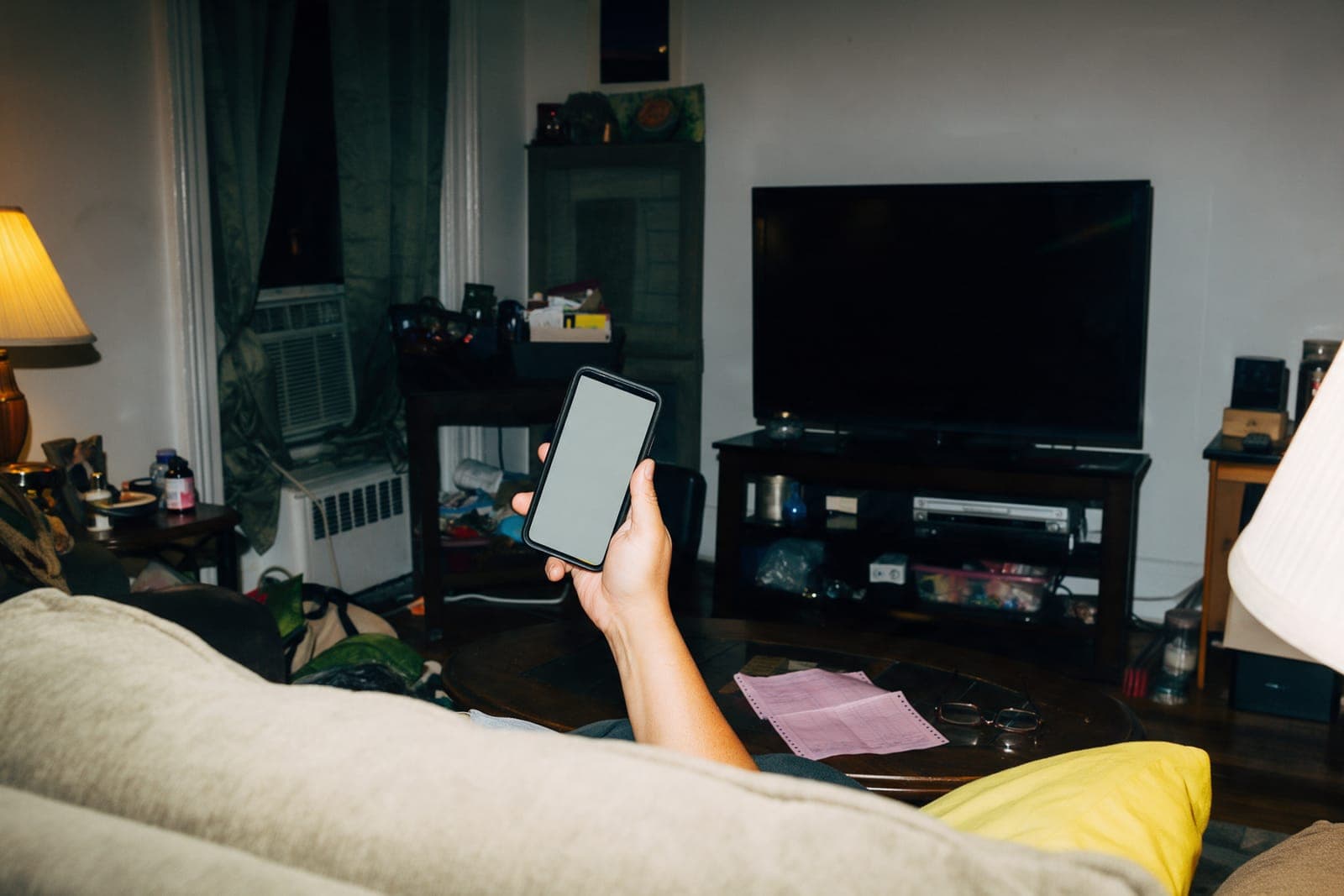 Over the back of a couch at night: a hand holds up a phone showing the signed quote, a pink paper estimate and reading glasses on the coffee table, a lamp glowing at the edge.