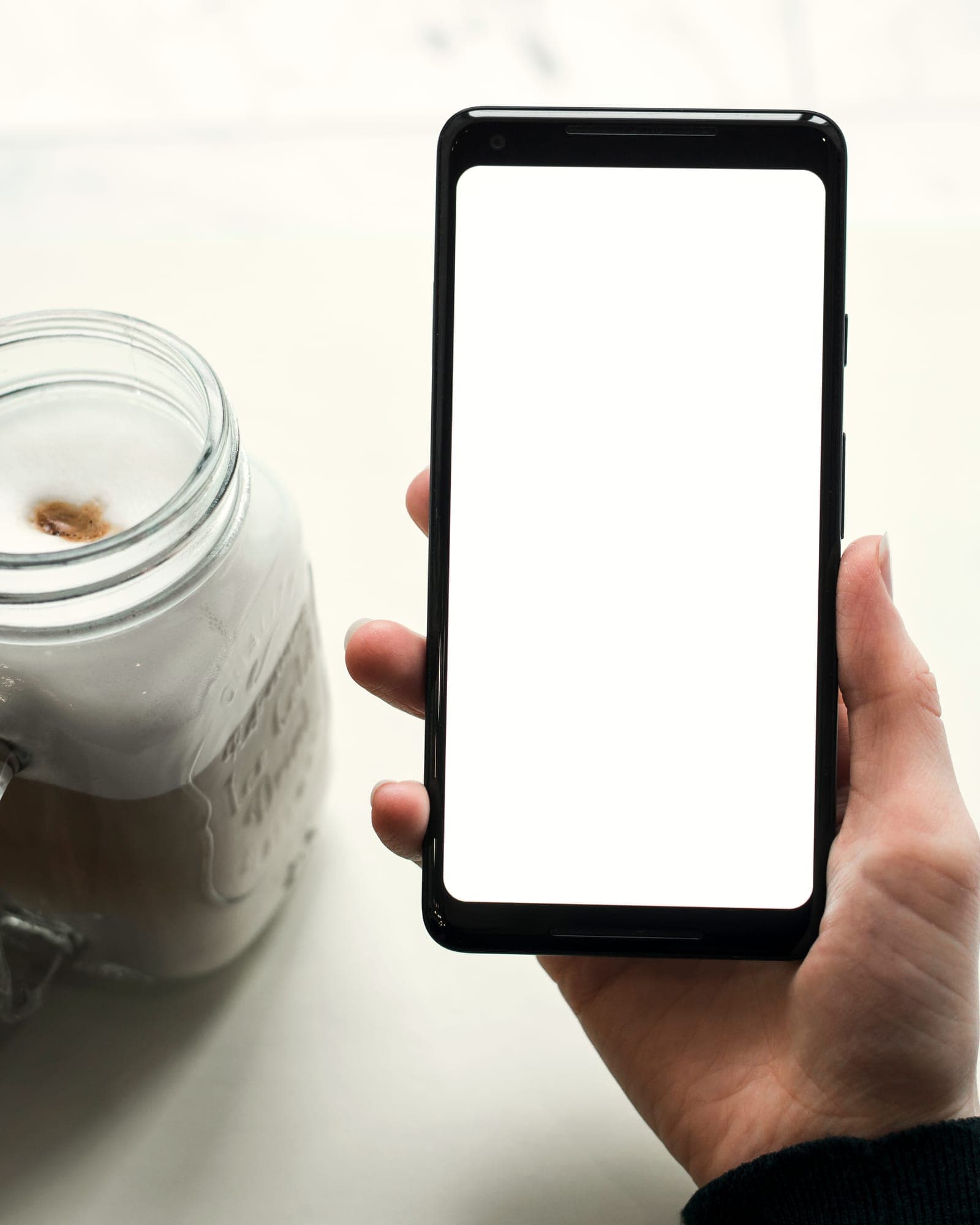 A hand holds a phone over a white marble counter, a latte in a glass jar beside it.