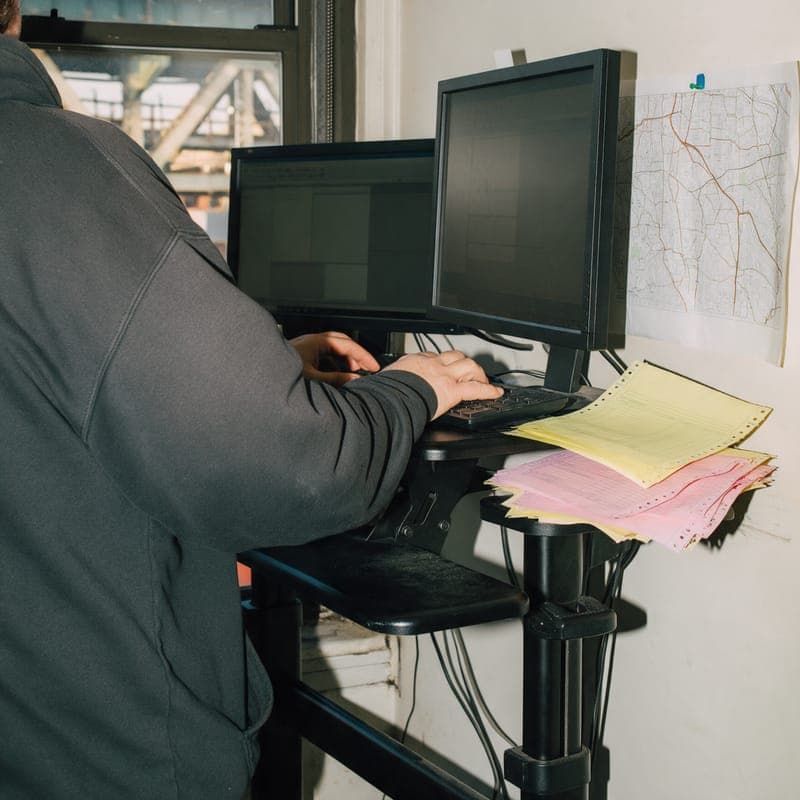A person at a standing desk by a factory window, an overhead train trestle outside and canary and pink copies beside the keyboard.