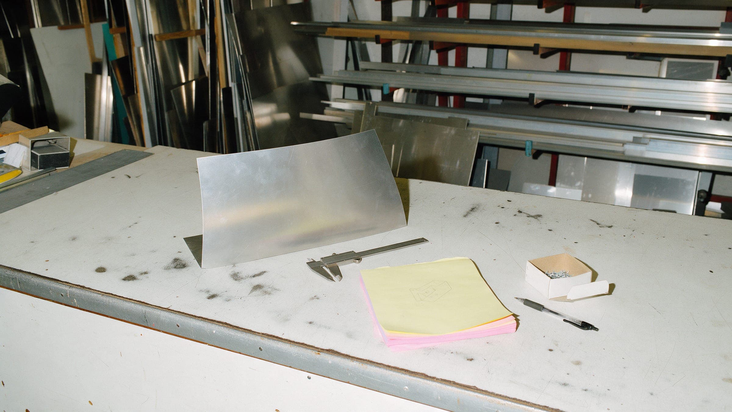 A fab shop counter with a bent aluminum sign blank, calipers and a sketched carbon pad.