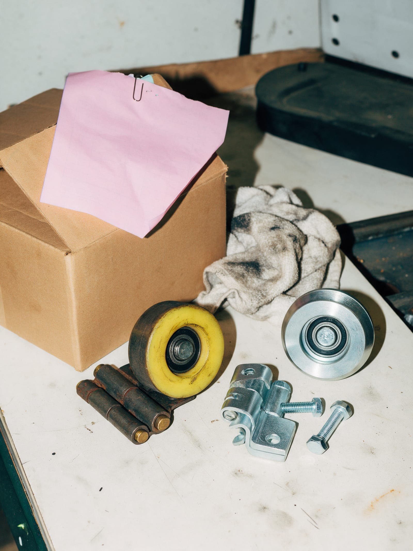 A worn garage door roller and hinge beside new parts on a workbench, a pink copy clipped to the carton.