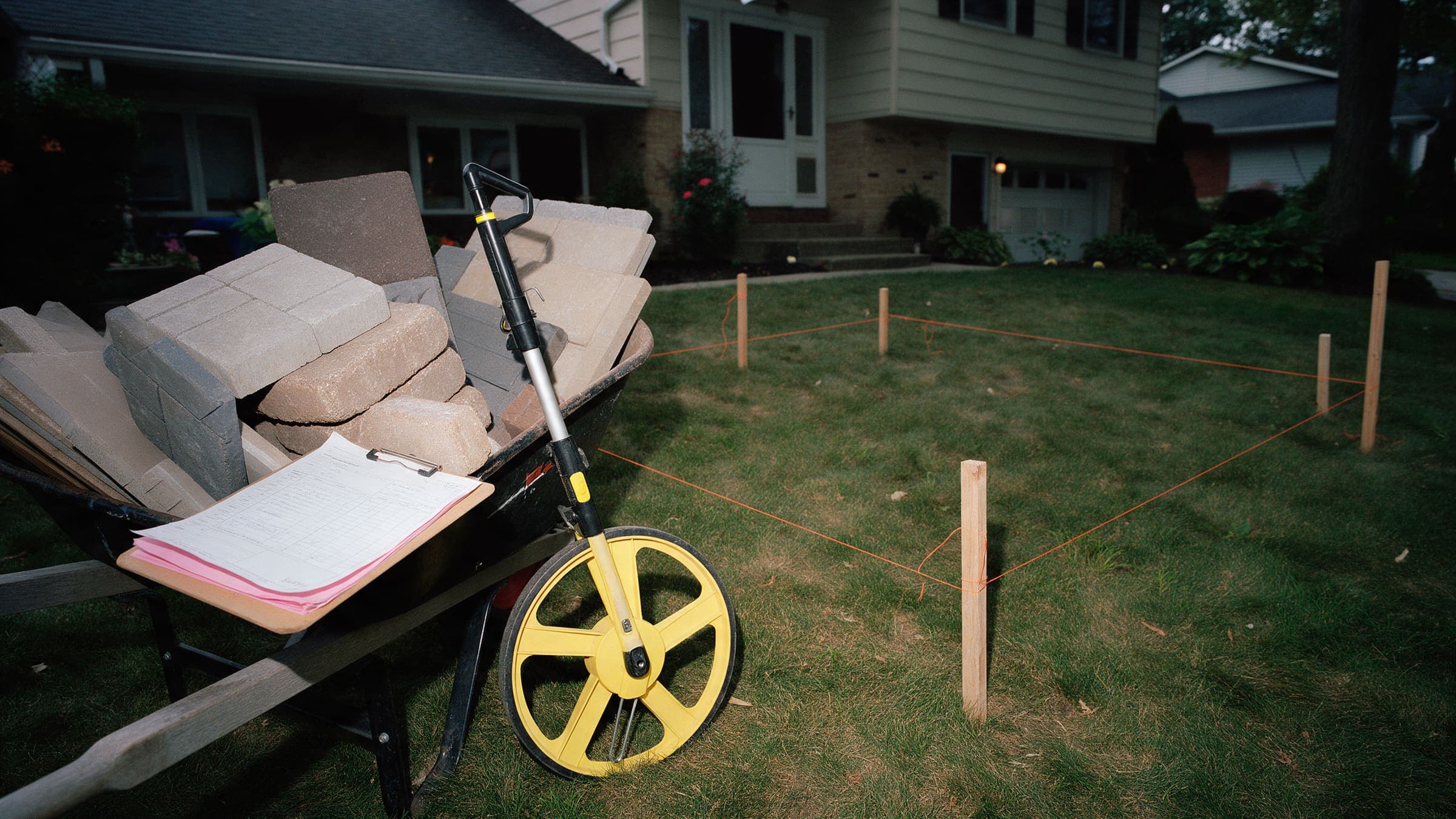 Paver samples in a wheelbarrow, a measuring wheel and a staked string outline for a new patio on a front lawn.