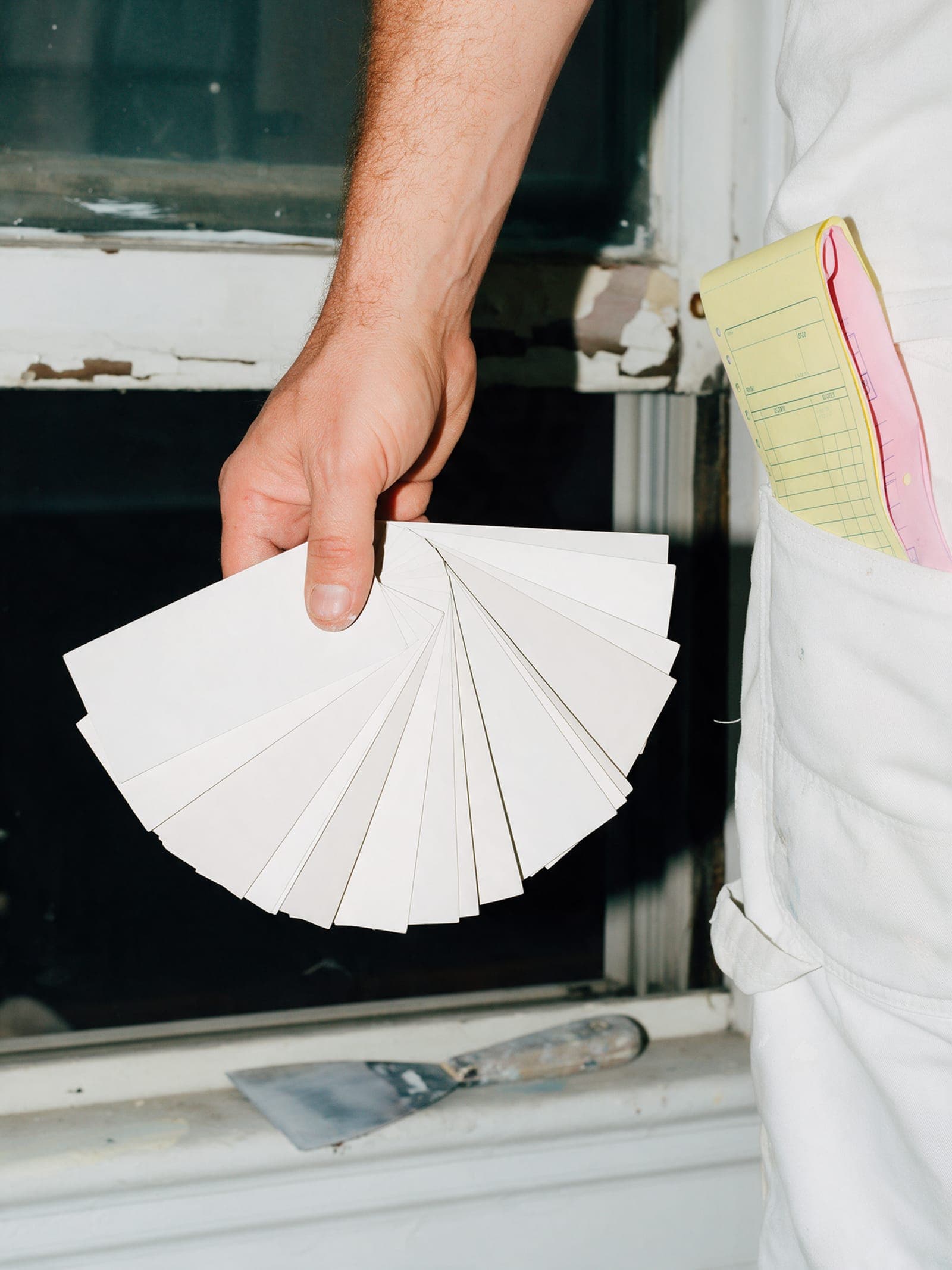 A painter's hand holding a fan of color chips by a peeling window casing, a canary copy in a back pocket.