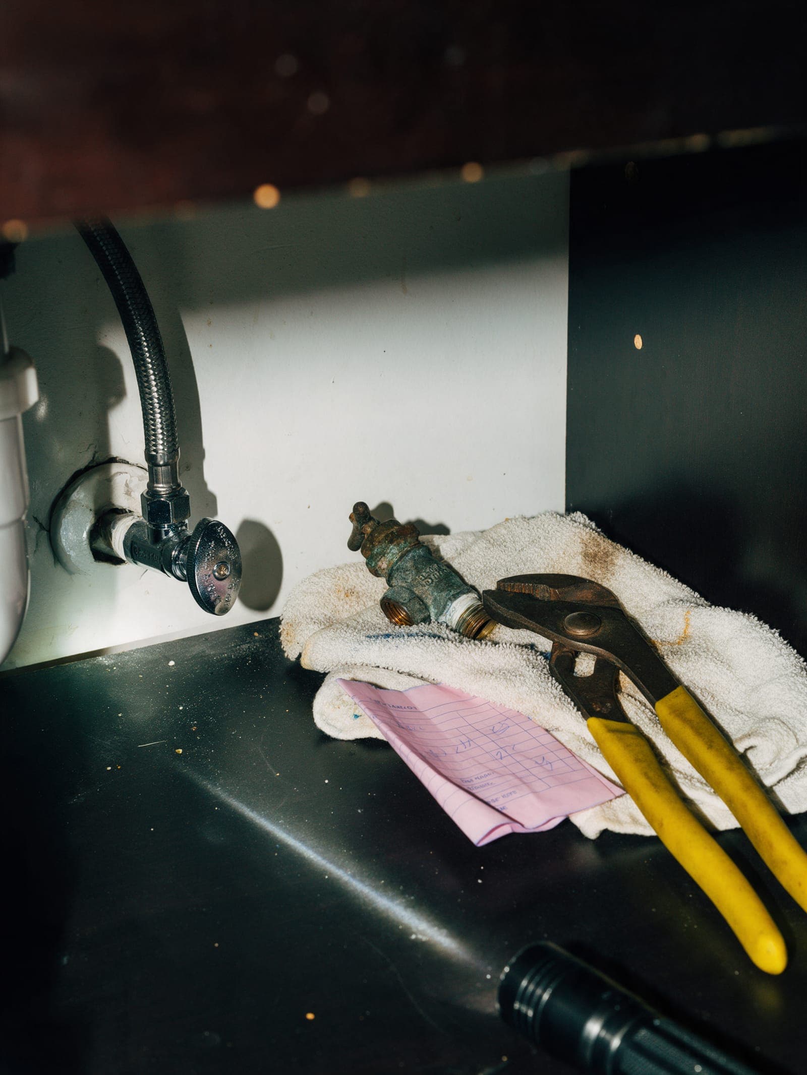 Under a bathroom vanity: a new chrome shutoff valve, the old corroded one on a towel, and a folded pink estimate copy.