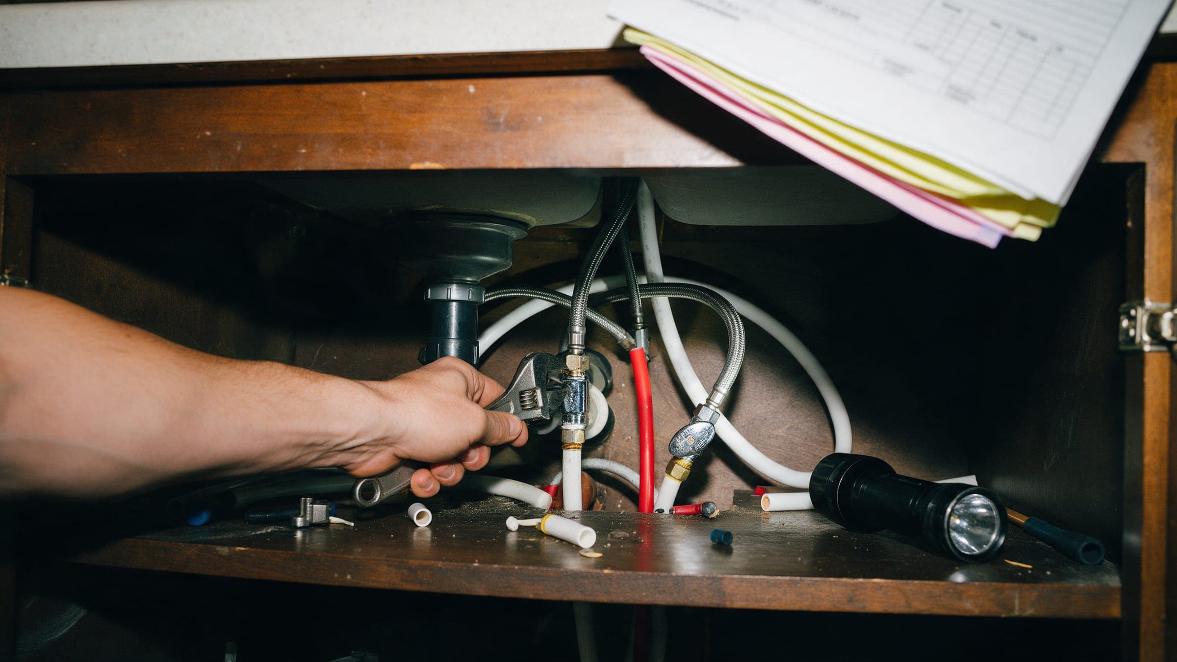 A plumber's forearm tightening a supply line under a kitchen sink.