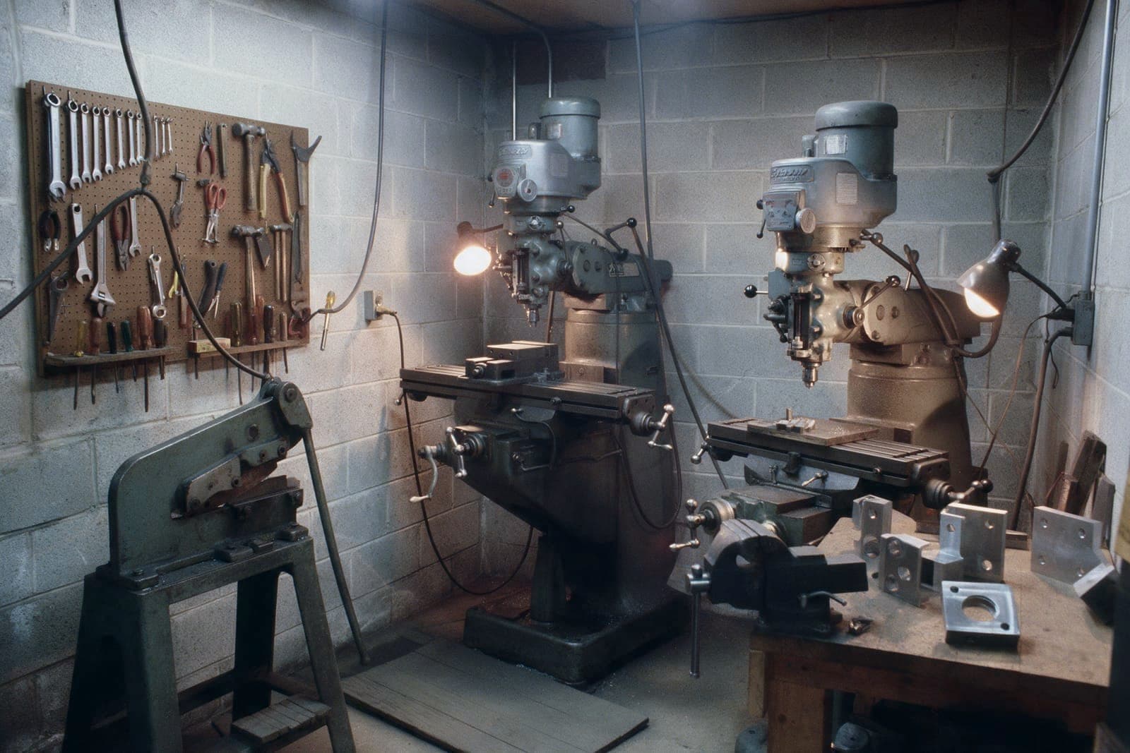A cramped tool-and-die bay with two manual mills, a shear and a pegboard of hand tools under warm work lamps