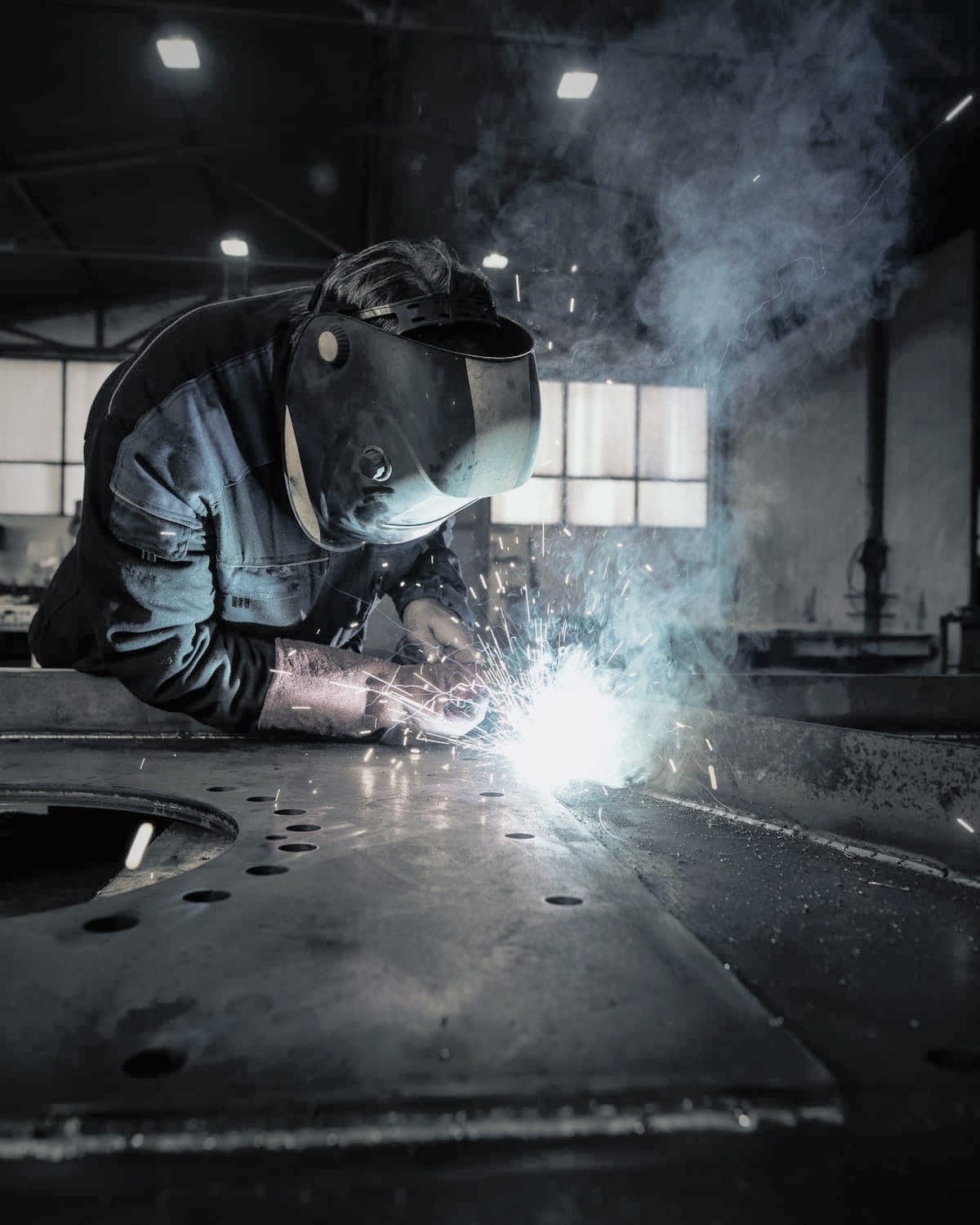 Welder in a helmet bent over a large plate, sparks and smoke rising from the arc