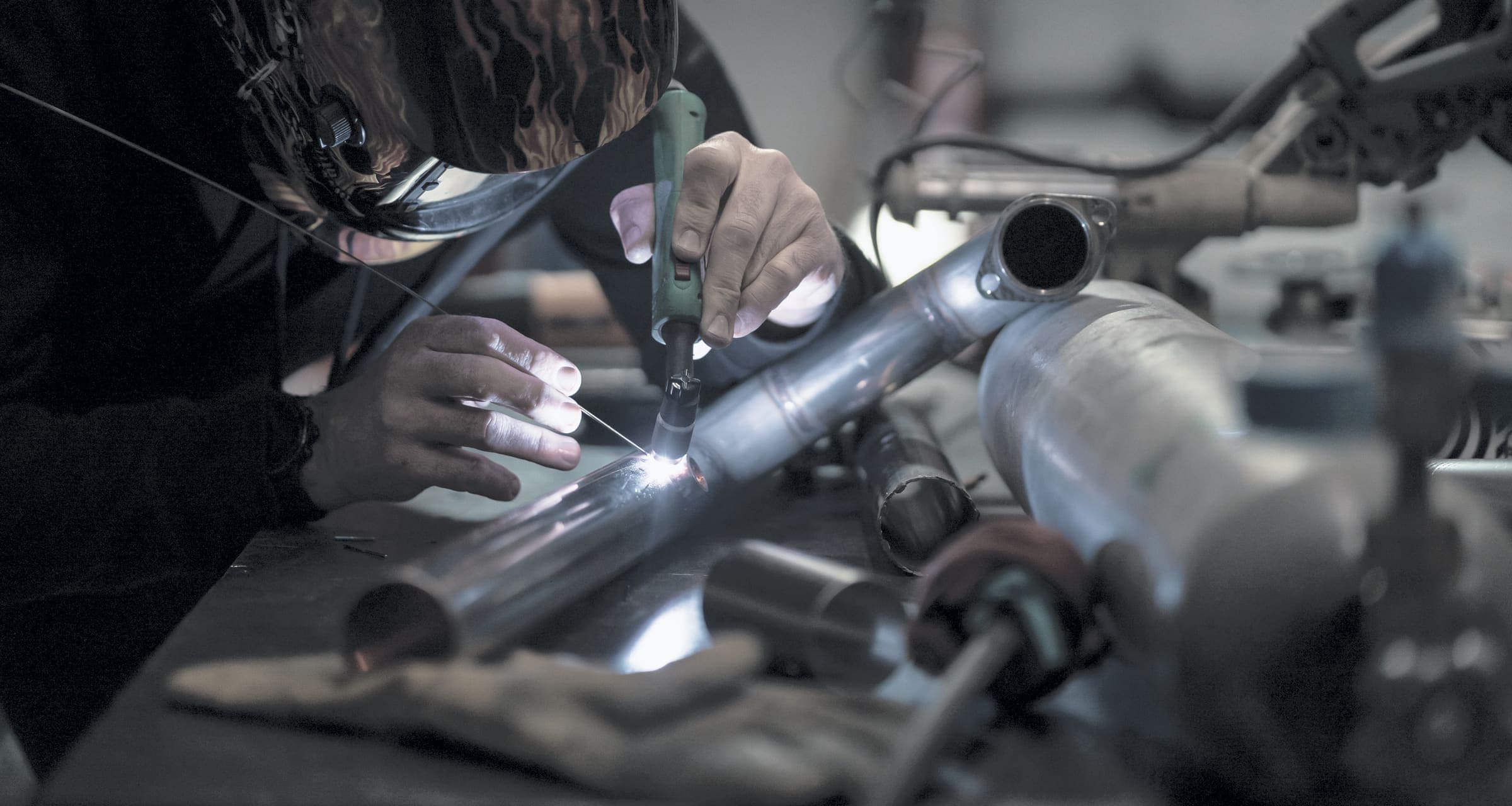 A TIG weld being laid on a stainless tube joint, torch and filler rod in hand, the welder behind a lowered helmet