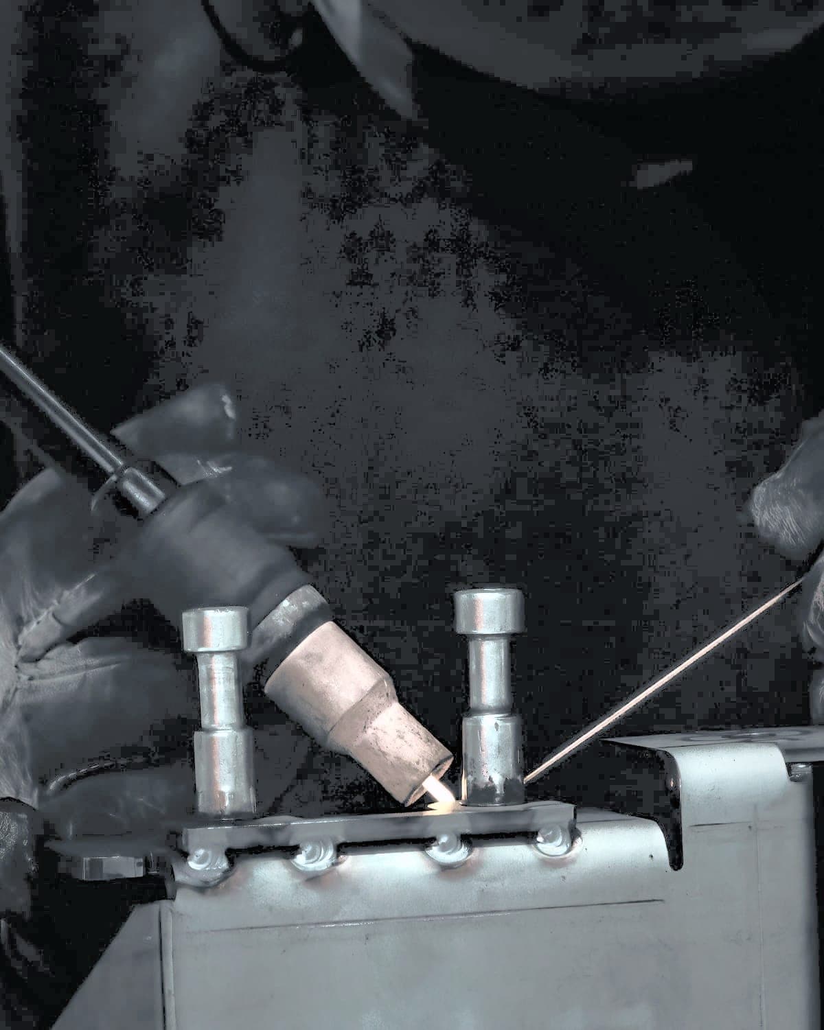 Gloved hands laying a TIG weld on a stainless part, the filler rod feeding in from the right