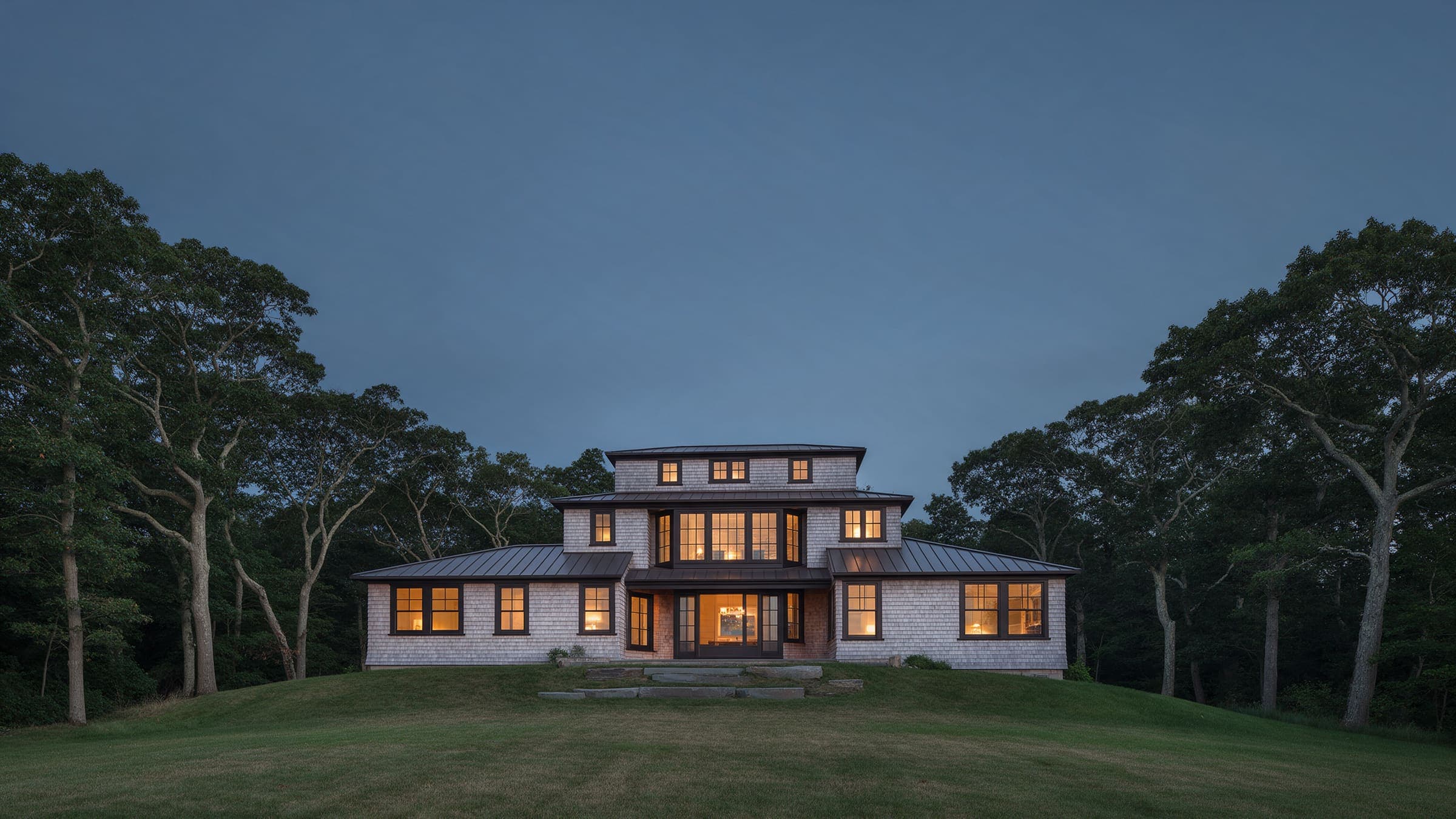 Bluff House from the bluff lawn at dusk, three stepped levels of cedar shingle with the living level lit.
