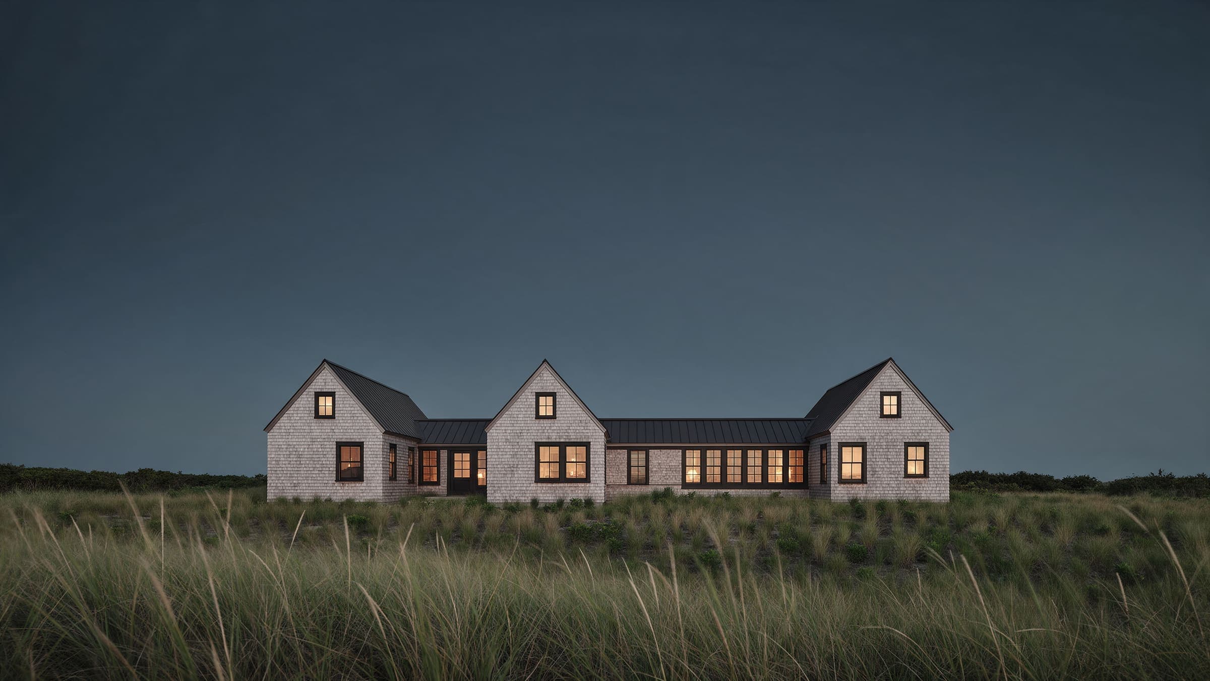 Dune Shingle House at dusk: three shingled gables joined by a low spine, windows lit, dune grass in the foreground.