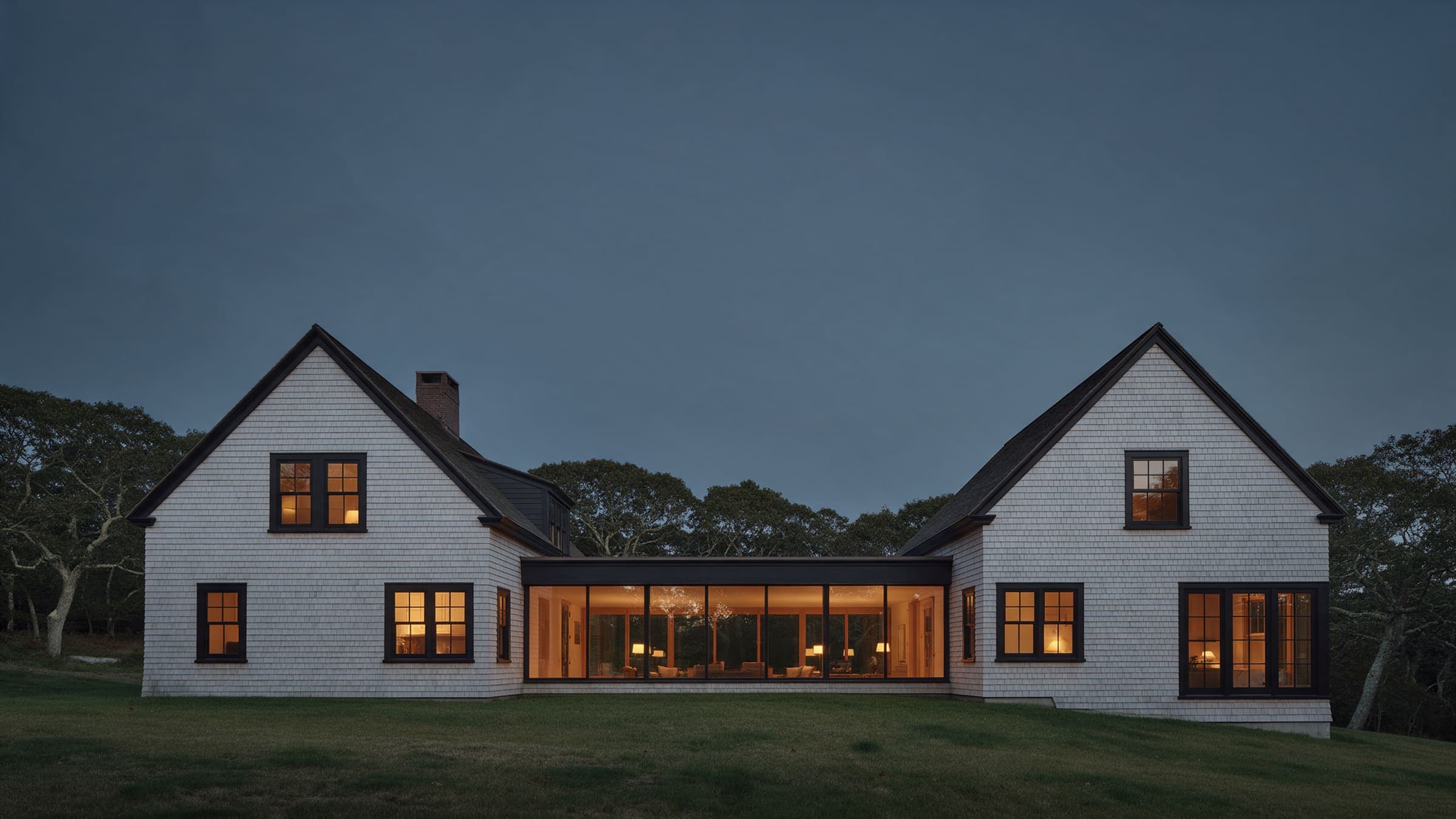 Gable Pair at dusk: two shingled 1920s gables joined by a long low glass hall, lit from inside.
