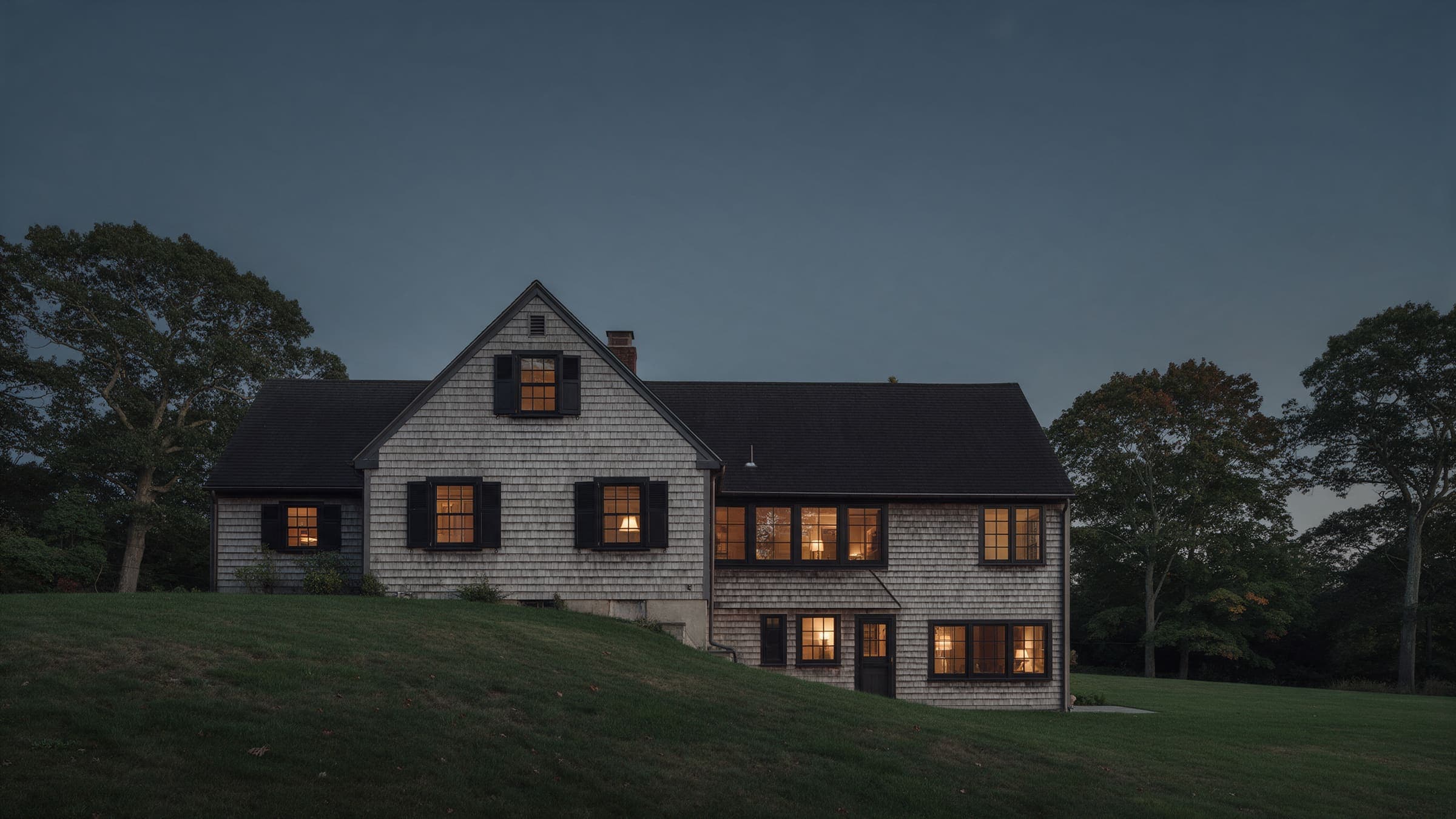 Harbor Hill Addition at dusk: a shingled cape with its new wing stepping down the lawn.