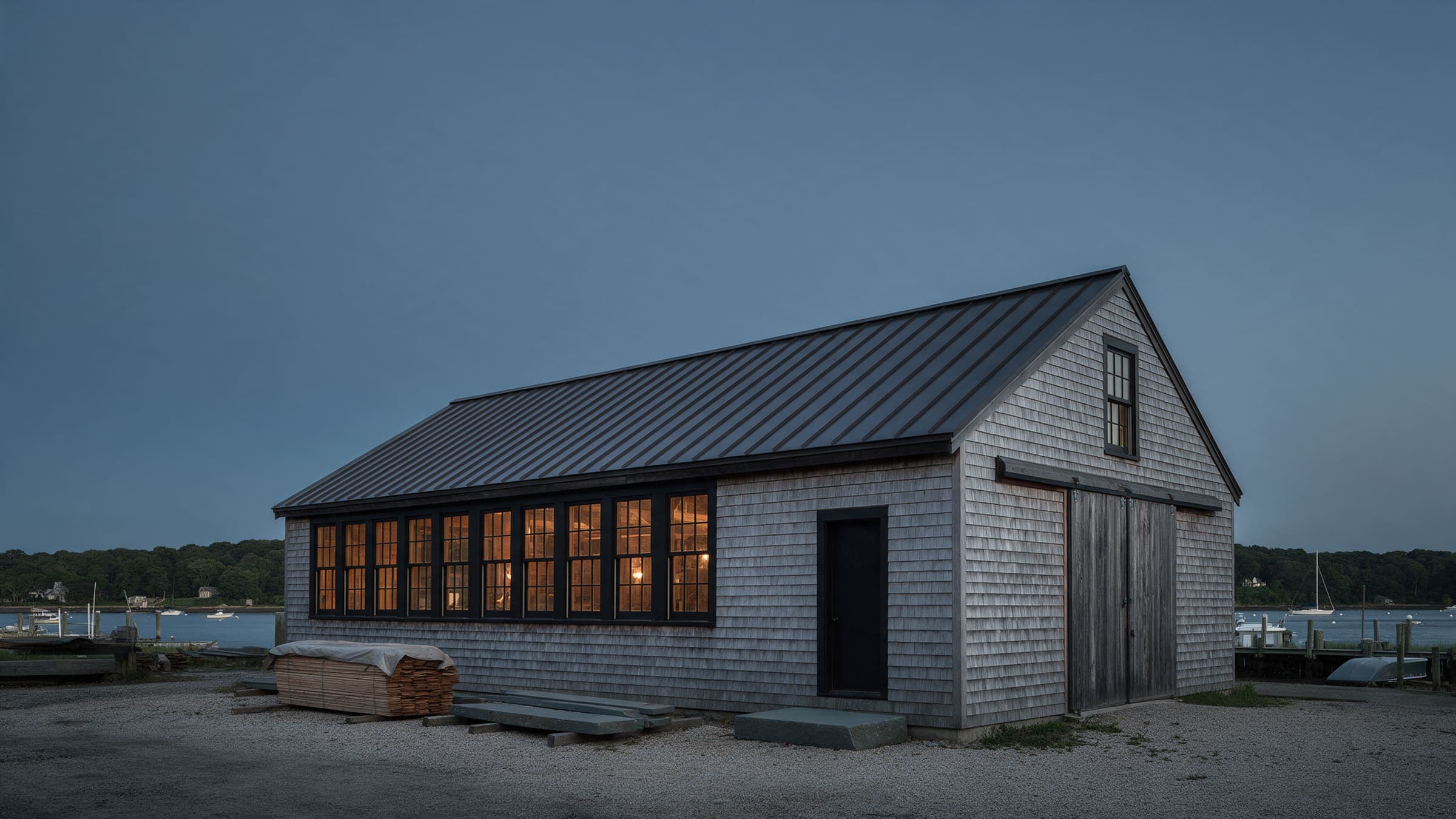 The studio at dusk: a long shingled boatyard shed with a row of lit windows, the harbor and moored boats behind.